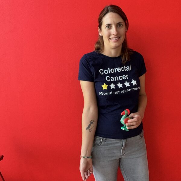 Johanna vor roter Wand mit Shirt. Aufdruck: Colorectal Cancer. 1 Stern. Would not recommend. und einem Papageientaucher-Tattoo